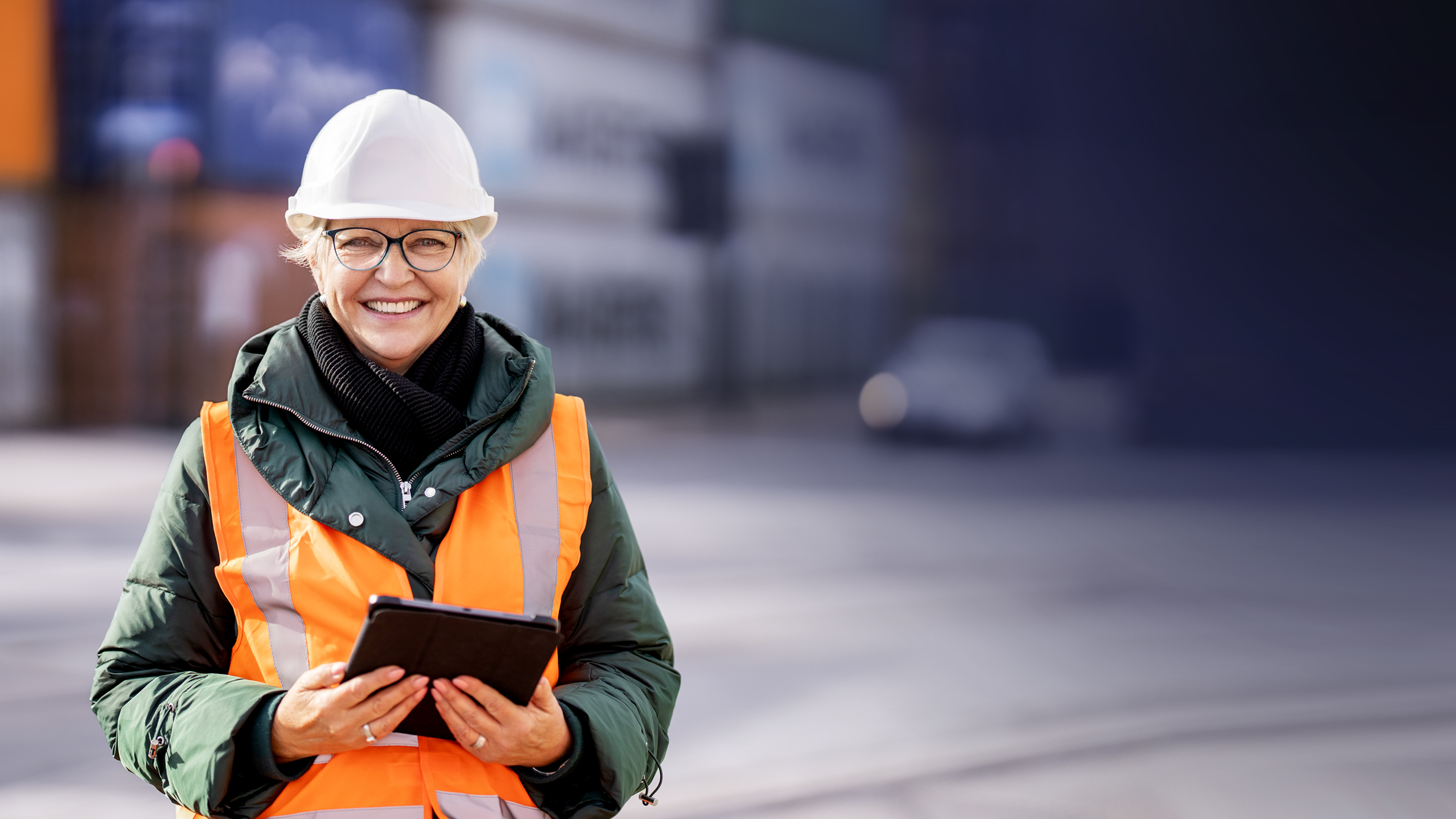 Herausforderungen Logistik Verkehr Frau in orangener West und mit Sicherheitshelm lächelt in die Kamera mit einem Tablet in der Hand